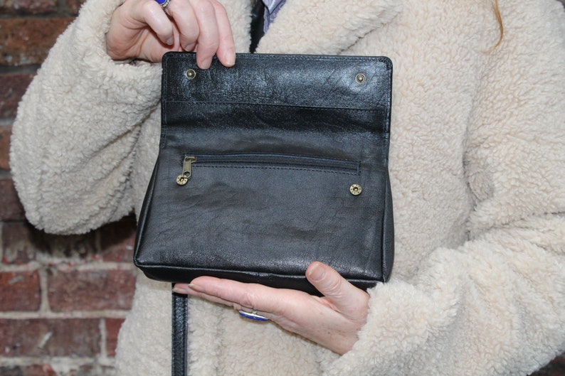 K&ouml;nnte beinhalten: Eine schwarze Leder-Umh&auml;ngetasche mit einer Rei&szlig;verschlusstasche und einem Klappverschluss. Die Tasche ist offen und das Innere ist sichtbar.