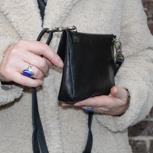 K&ouml;nnte beinhalten: Eine schwarze Leder-Umh&auml;ngetasche mit einem langen Riemen. Die Tasche wird mit einer Klappe geschlossen und hat einen silbernen Verschluss.
