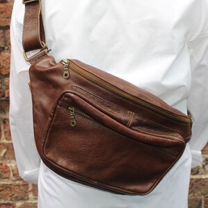 K&ouml;nnte beinhalten: Eine braune Leder-Bauchtasche mit einem Rei&szlig;verschlussfach und einem langen verstellbaren Riemen. Die Tasche wird &uuml;ber der Schulter getragen.