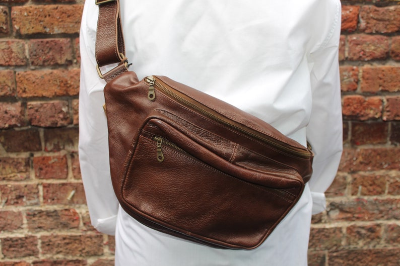 K&ouml;nnte beinhalten: Eine braune Leder-Bauchtasche mit einem Rei&szlig;verschlussfach und einem langen verstellbaren Riemen. Die Tasche wird &uuml;ber der Schulter getragen.