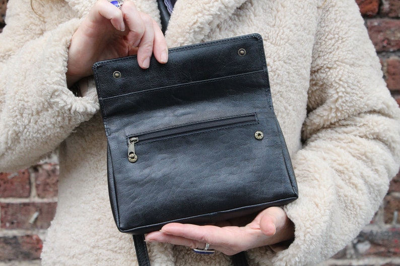 K&ouml;nnte beinhalten: Eine schwarze Lederhandtasche mit einem Rei&szlig;verschlussfach und einem Klappverschluss. Die Tasche ist offen und das Innere ist sichtbar.