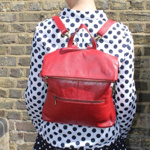 May include: A red leather backpack with a flap and a front zip pocket. The bag has adjustable straps and a top handle. The backpack is shown against a brick wall.