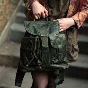 May include: A dark green leather rucksack with two large front pockets and a buckle closure. The rucksack has a drawstring closure at the top and adjustable straps.