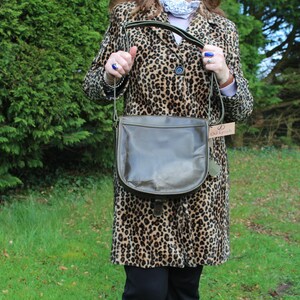 May include: A brown leather satchel bag with a green strap and a brown leather flap. The bag is being held by a person wearing a leopard print coat.