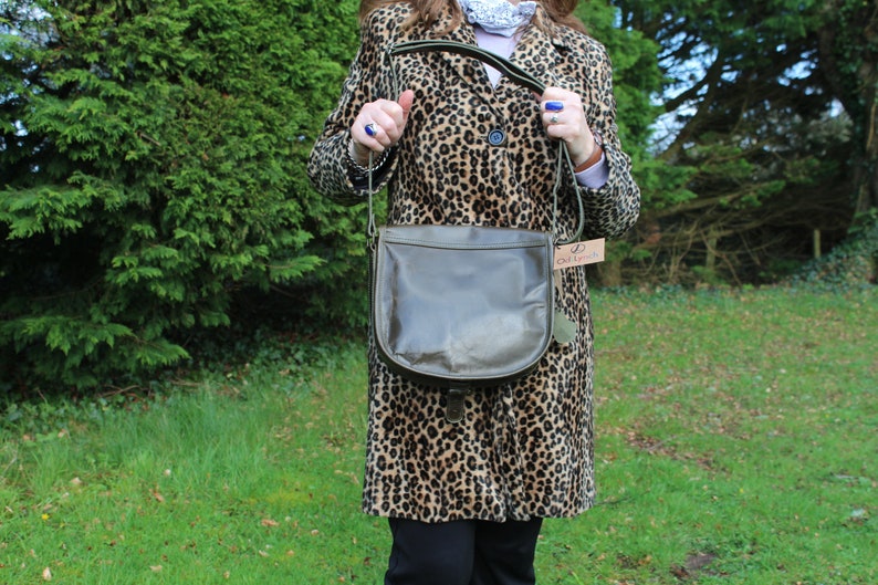 May include: A brown leather satchel bag with a green strap and a brown leather flap. The bag is being held by a person wearing a leopard print coat.