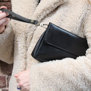 K&ouml;nnte beinhalten: Eine schwarze Leder-Clutch mit einem kurzen Riemen. Die Tasche wird von einer Person gehalten, die eine cremefarbene, flauschige Jacke tr&auml;gt.