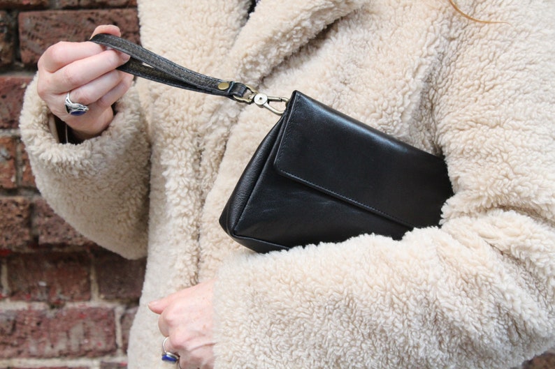 K&ouml;nnte beinhalten: Eine schwarze Leder-Clutch mit einem kurzen Riemen. Die Tasche wird von einer Person gehalten, die eine cremefarbene, flauschige Jacke tr&auml;gt.