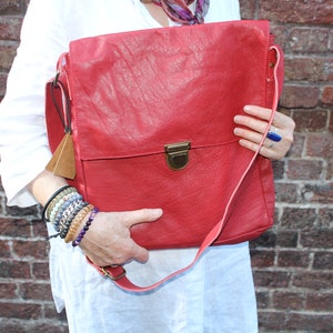 Puede incluir: Un bolso mensajero de cuero rojo con cierre de solapa y correa para el hombro. El bolso tiene una superficie texturizada y un cierre de color latón. Una pequeña etiqueta de cuero cuelga del lateral. El bolso se sostiene frente a una pared de ladrillos.