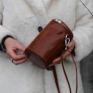 May include: A cylindrical brown leather bag with a silver-tone clasp and a matching strap. The bag is held by a person wearing a white coat.