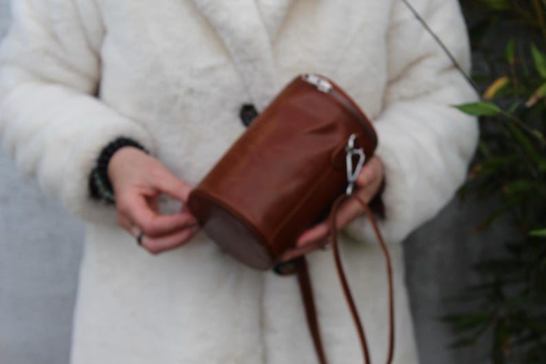 May include: A cylindrical brown leather bag with a silver-tone clasp and a matching strap. The bag is held by a person wearing a white coat.