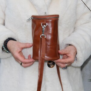May include: A cylindrical, brown leather handbag with a zipper closure and a long, thin strap. The bag has a silver-colored clasp and a small, round button on the bottom. The bag is held in front of a white coat.