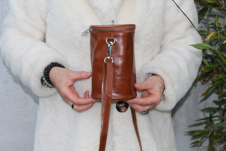 May include: A cylindrical, brown leather handbag with a zipper closure and a long, thin strap. The bag has a silver-colored clasp and a small, round button on the bottom. The bag is held in front of a white coat.