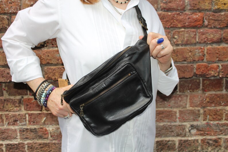 May include: Black leather fanny pack with a front zippered pocket and a top zippered compartment. The bag has a black adjustable strap and gold-colored zippers. The bag is worn across the body.