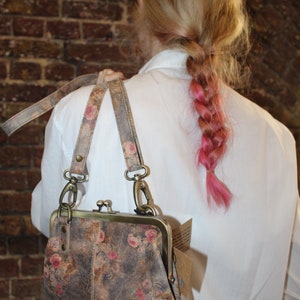 May include: A floral patterned leather purse with a brass clasp and a long strap. The purse is worn by a person with pink hair.