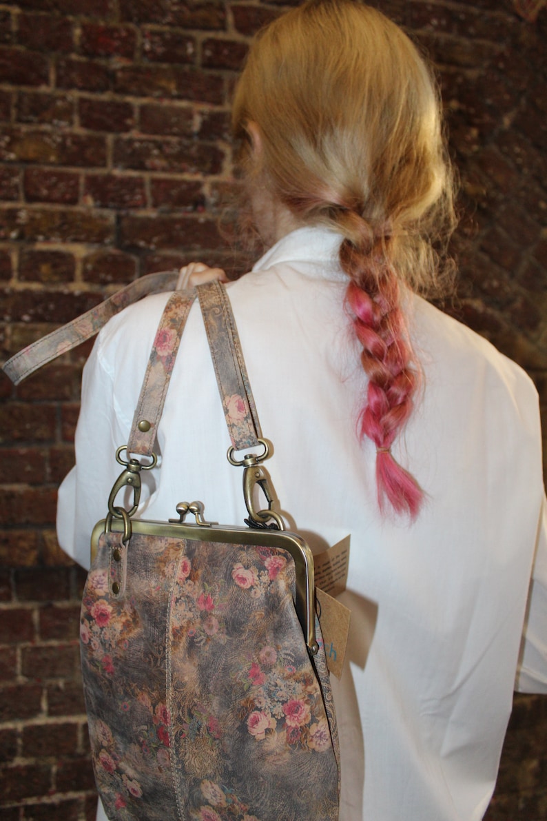 May include: A floral patterned leather purse with a brass clasp and a long strap. The purse is worn by a person with pink hair.