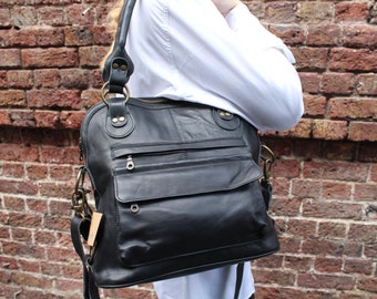 Puede incluir: Bolso de hombro de cuero negro con múltiples bolsillos y herrajes de color latón. El bolso tiene un asa superior y una correa para el hombro desmontable. El bolso se muestra sobre un fondo de pared de ladrillo.