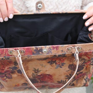 May include: A vintage-style handbag with a floral pattern in shades of red, brown, and beige. The bag has a black interior with pockets and light brown handles. The clasp is silver-toned.