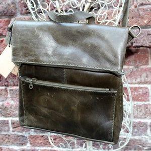 May include: An olive green leather backpack with a flap closure and a front zipped pocket. The bag features a top handle and adjustable shoulder straps. A small tag hangs from the top flap. The bag is displayed against a brick wall.