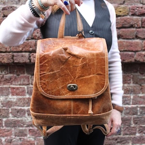 Puede incluir: Una mochila de cuero marrón con cierre de solapa y cordón de ajuste. La mochila tiene un aspecto desgastado y está hecha de cuero suave y flexible.