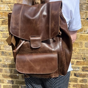 May include: A brown leather rucksack with a flap closure and a front pocket. The bag has a drawstring closure and adjustable straps. The leather has a distressed appearance. The rucksack is designed for carrying books, laptops, and other essentials.