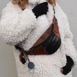 May include: A brown and black leather fanny pack with a zipper closure. The fanny pack is worn by a person wearing a white fluffy jacket.