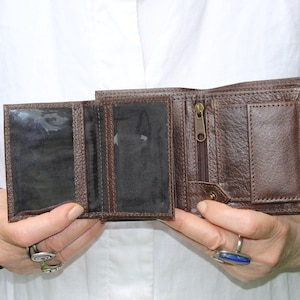 May include: A dark brown leather bifold wallet featuring a textured surface, two clear plastic ID windows on the left side, a vertical zippered compartment with a metal pull, and a small flap closure with a snap button on the right side.