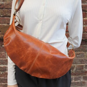 May include: A brown leather crossbody bag with a curved shape and a long strap. The bag is worn over the shoulder of a person wearing a white shirt.