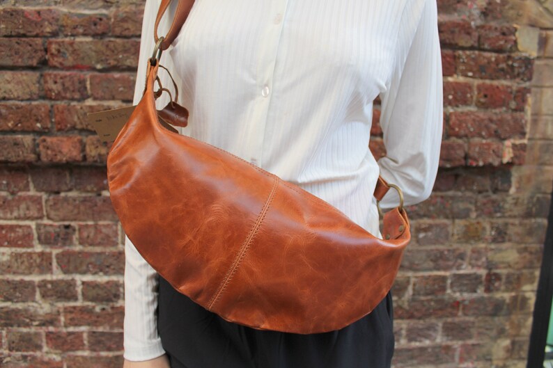 May include: A brown leather crossbody bag with a curved shape and a long strap. The bag is worn over the shoulder of a person wearing a white shirt.
