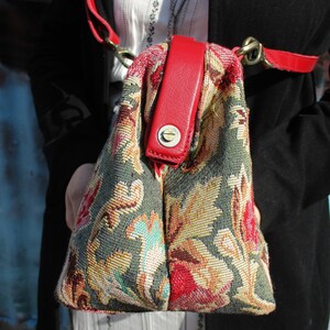 May include: A floral tapestry handbag with a red leather strap and a brass clasp. The bag has a green, beige, and red floral pattern.