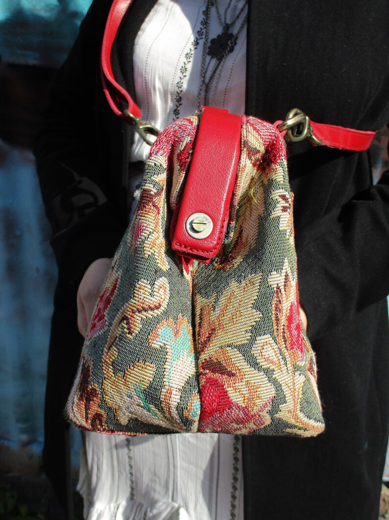 May include: A floral tapestry handbag with a red leather strap and a brass clasp. The bag has a green, beige, and red floral pattern.