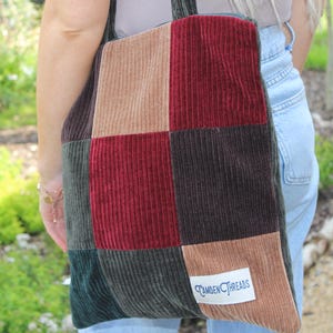 May include: A patchwork corduroy tote bag in shades of brown, burgundy, green, and tan. The bag has a rectangular shape with a label that reads "Camden Threads". The bag is carried over the shoulder.