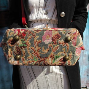 May include: A tapestry-style handbag with a floral pattern in shades of red, green, and gold. The bag has four metal studs on the bottom.