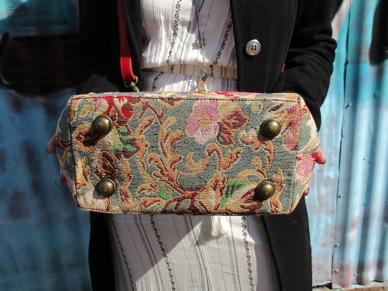 May include: A tapestry-style handbag with a floral pattern in shades of red, green, and gold. The bag has four metal studs on the bottom.