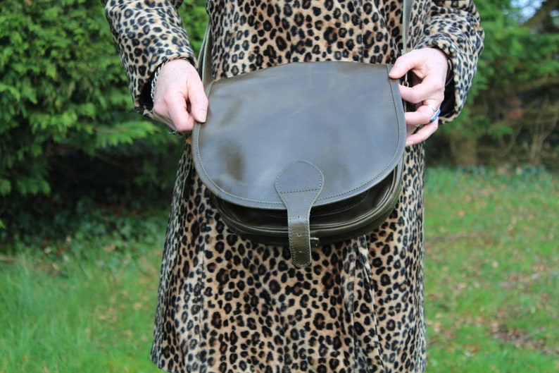 May include: A dark green leather satchel bag with a flap closure and a buckle. The bag is worn by a person wearing a leopard print coat.