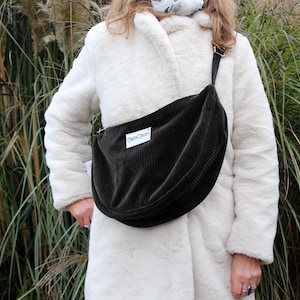 Op de afbeelding: Een zwarte corduroy crossbody tas met een gebogen vorm en een lange verstelbare riem. De tas heeft een rechthoekig wit label met tekst. De persoon draagt een witte imitatiebontjas.