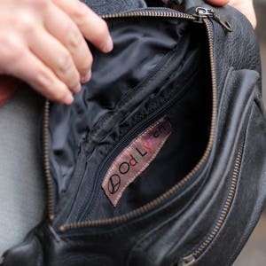 May include: A black leather fanny pack with a zipper closure and a small pocket on the front. The inside of the bag is lined with black fabric and has a small pocket with a pink label that says "Oddlyfern".