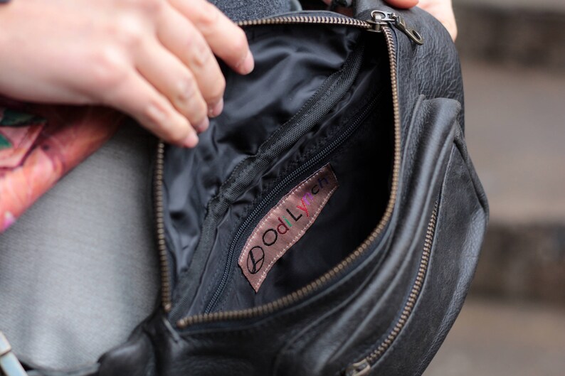 May include: A black leather fanny pack with a zipper closure and a small pocket on the front. The inside of the bag is lined with black fabric and has a small pocket with a pink label that says "Oddlyfern".