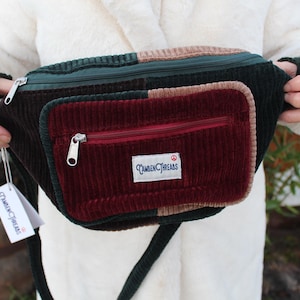 May include: A multi-colored corduroy fanny pack with a zippered main compartment and a smaller front pocket. The pack features a dark green, brown, and burgundy color scheme, with a white tag that reads "Camden Threads".