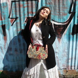 May include: A woman wearing a white dress with a black jacket and a floral patterned handbag with a red strap. She is standing in front of a blue and white graffiti wall.