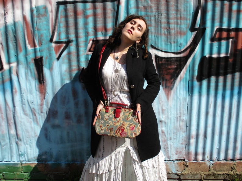 May include: A woman wearing a white dress with a black jacket and a floral patterned handbag with a red strap. She is standing in front of a blue and white graffiti wall.