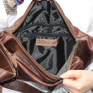 K&ouml;nnte beinhalten: Eine braune Leder-Bauchtasche mit einem Rei&szlig;verschlussfach und einer kleineren Rei&szlig;verschlusstasche auf der Vorderseite. Das Innere der Tasche ist mit schwarzem Stoff ausgekleidet und tr&auml;gt ein Etikett mit der Aufschrift "Odd Lynch".