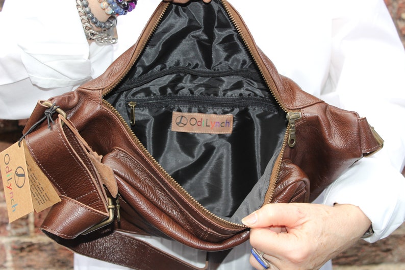 K&ouml;nnte beinhalten: Eine braune Leder-Bauchtasche mit einem Rei&szlig;verschlussfach und einer kleineren Rei&szlig;verschlusstasche auf der Vorderseite. Das Innere der Tasche ist mit schwarzem Stoff ausgekleidet und tr&auml;gt ein Etikett mit der Aufschrift "Odd Lynch".