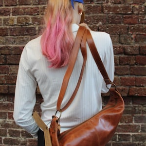May include: A woman wearing a white shirt with a brown leather shoulder bag. The bag has a long strap and a flap closure. The woman is wearing a blue baseball cap and has pink hair.