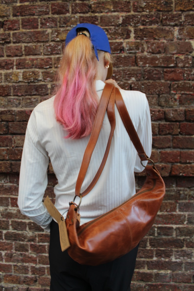 May include: A woman wearing a white shirt with a brown leather shoulder bag. The bag has a long strap and a flap closure. The woman is wearing a blue baseball cap and has pink hair.