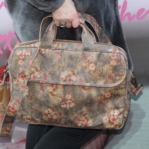 May include: A floral-patterned satchel bag with a shoulder strap and top handle. The bag features a muted grey background with pink and red rose accents. It has a front flap pocket and a tag attached to the strap.