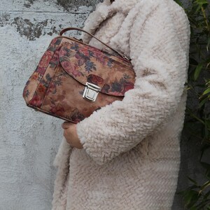 May include: A brown handbag featuring a floral print, a silver clasp, and a top handle. The bag is held against a cream-coloured, textured coat. The floral pattern includes red, pink, and blue flowers.