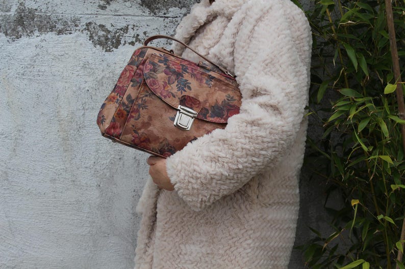 May include: A brown handbag featuring a floral print, a silver clasp, and a top handle. The bag is held against a cream-coloured, textured coat. The floral pattern includes red, pink, and blue flowers.