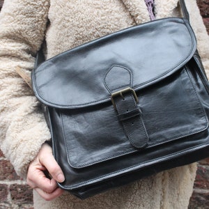 May include: Black leather messenger bag with a flap closure and a buckle. The bag has a front pocket and a shoulder strap. The bag is being held in front of a brick wall.