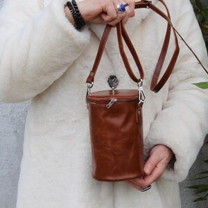 May include: A brown leather cylindrical crossbody bag with a zipper closure and a long, adjustable strap. The bag is held by a person wearing a white coat. The bag's hardware is silver-colored.