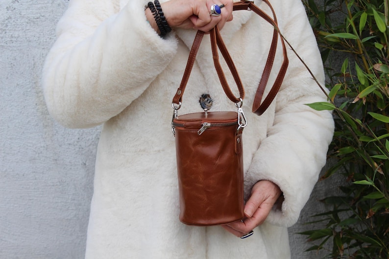 May include: A brown leather cylindrical crossbody bag with a zipper closure and a long, adjustable strap. The bag is held by a person wearing a white coat. The bag's hardware is silver-colored.
